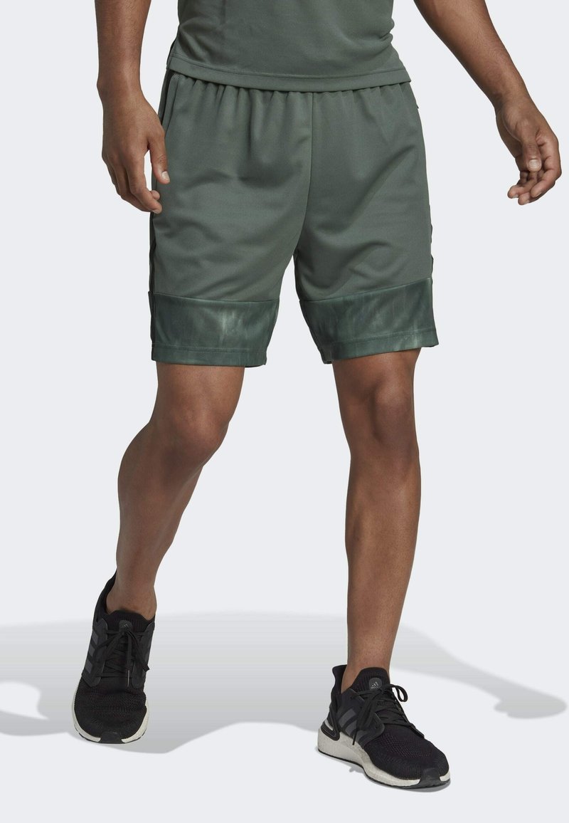 Man i mörkgröna träningsshorts med svarta detaljer och svarta löparskor, stående med ett ben framåt mot en vit bakgrund.