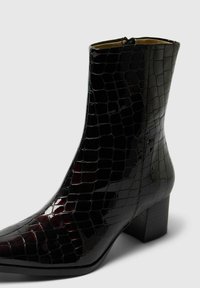 Melna patent ādas potīšu zābaku pāris ar krokodila tekstūru, ar rāvējslēdzēju un bloku papēdi. Iezīmējas ar spīdīgu apdari un smailu purngalu.