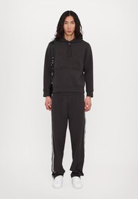Um hoodie cinza escuro e calças de treino combinando, ambos com um logótipo texturizado, apresentando riscas brancas à lateral. Ténis brancos completam o visual.