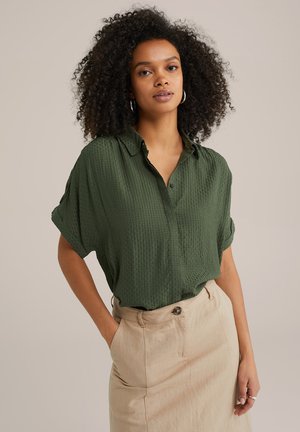 Grünes, strukturiertes Kurzarmhemd mit einem Button-down-Kragen, kombiniert mit einem hoch taillierten beigen Rock, der einen einzelnen vorderen Knopf hat.