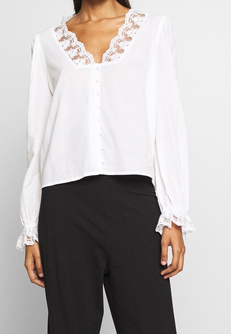 Chemisier blanc avec une bordure en dentelle autour de l'encolure et des manches, présentant un devant à boutons et une silhouette ample. Un pantalon noir uni est porté en dessous.