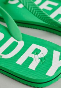 Groene, textuur aangepaste teenslippers met grote witte letters die "DRY" spellen, zwarte rand aan de zool en een chevronpatroon op de bandjes, geplaatst op een grijze ondergrond.