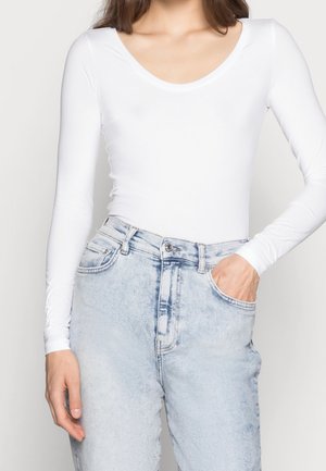 Femme portant un haut blanc moulant à manches longues et un jean taille haute bleu clair, une main dans la poche.