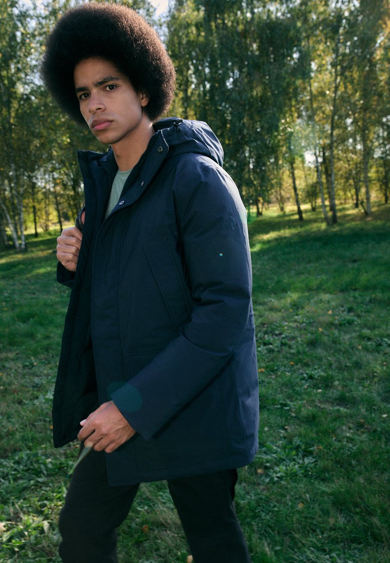 Navy blue insulated jacket with a hood, featuring a zip closure and side pockets, worn over a light shirt in an outdoor setting.