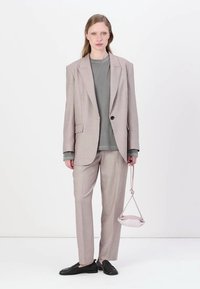 Blazer e calças combinando, levemente padronizados, em bege e castanho, emparelhados com uma camisa cinza e uma pequena mala rosa. Usando mocassins pretos.