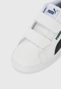 Basket de sport blanche avec deux bandes Velcro, accent noir sur le côté, matériau synthétique lisse et semelle en caoutchouc. Logo vert sur la languette.