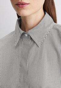 Chemise à rayures noires et blanches avec col pointu, fermeture à bouton et texture en coton lisse, mettant l'accent sur les lignes verticales.