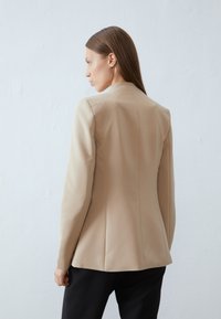 Blazer beige ajustado com ombros estruturados, costuras nas costas e um acabamento limpo. Tecido suave, sem fechos visíveis. Usado sobre calças pretas.
