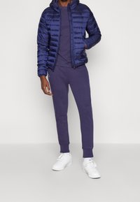 Marine-blauwe pufferjack met horizontale stiksels, gecombineerd met een paarse sweatshirt en bijpassende joggingsbroek. Witte sneakers maken de look compleet.