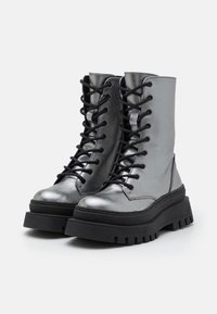 Botas de combate metálicas plateadas con cordones negros, diseño de tobillo alto, suela gruesa negra y una textura suave. Presenta costuras robustas.