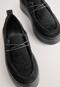 Svarte slip-on sko i ruskind med en tyk sort plateau-sål, der har grå snørebånd og rund tå. Materialet har tekstur, der giver dybde.