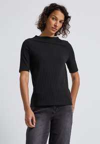 Kvinde med krøllet hår iført en sort ribbet kortærmet top og mørkegrå jeans, stående imod en ensfarvet lys baggrund.