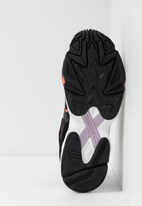 Svart och vit sneakersula med strukturerat greppmönster, med en orange detalj och en lila torsionsplatta för stabilitet.