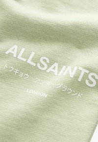 Svetlo zelena tkanina vsebuje belo besedilo z napisom "ALLSAINTS," dodatno besedilo v japonščini in "LONDON" na dnu. Gladka tekstura.