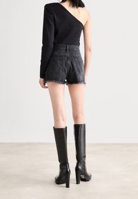 Mujer de espaldas que lleva una blusa negra de un solo hombro, pantalones cortos de mezclilla negra de talle alto con bordes deshilachados, y botas altas negras de tacón.