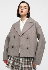 Νεαρό άτομο που φοράει ένα μπεζ παλτό διπλής σειράς κουμπιών πάνω από ένα λευκό πουκάμισο και καρό πλισέ καφέ φούστα, στέκεται μπροστά σε απλό φόντο.