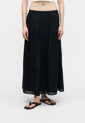 Nő fekete, hosszú, réteges szoknyában és fekete papucsban, egyszínű fehér háttér előtt áll.