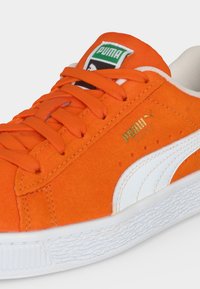 Φωτεινό πορτοκαλί αθλητικό παπούτσι Puma Suede με λευκή λωρίδα λογότυπου, πορτοκαλί κορδόνια, ανάγλυφη λευκή σόλα και πράσινη ετικέτα Puma στη γλώσσα.