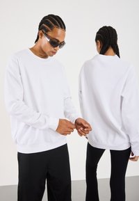 Two people with braided hair wearing white sweatshirts; one faces forward adjusting sleeves, the other shows the back of the sweatshirt.