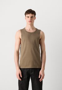 Lys beige ermeløs tank top laget av bomull, med rund hals og en subtil logo på brystet. Kombinert med mørke jeans.