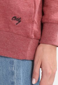 Lähedalt pilt käest punase sviitri varruka kõrval, millel on must "Ainult" tikand, ja siniste denimpükste kohal.