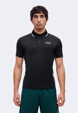 Młody mężczyzna o ciemnych kręconych włosach, ubrany w czarną koszulkę polo EA7 Emporio Armani i ciemnozielone szorty, stoi twarzą do przodu.