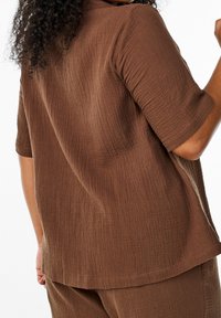 Braun strukturierter Oberteil mit kurzen Ärmeln, im entspannten Schnitt und mit dezenten Nähten. Der Stoff hat ein gerüschtes Aussehen und einen weichen Fall.