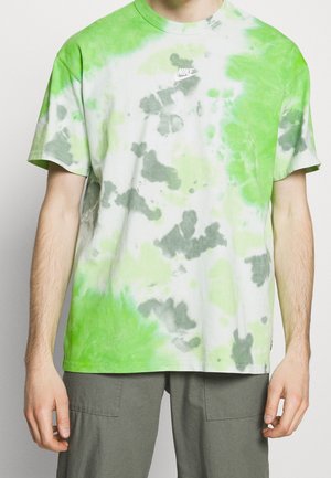 Person iført en grøn, hvid og grå tie-dye Nike T-shirt med korte ærmer og olivengrønne bukser, stående mod en ensfarvet baggrund.
