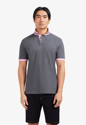 Homme portant un polo gris avec un col et des bords de manches roses, associé à un short noir, debout devant un fond blanc.