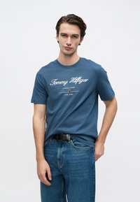 Μπλε βαμβακερό μπλουζάκι με λευκή λογότυπη επιγραφή και διακοσμητικά στοιχεία. Λαιμόκοψη τύπου crew, κοντά μανίκια, συνδυασμένο με μπλε τζιν παντελόνι.