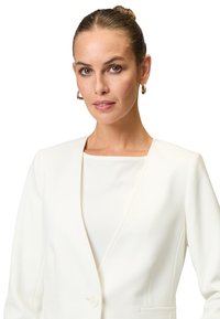Blazer bianco con un design strutturato, caratterizzato da una profonda scollatura a V, chiusura con singolo bottone e tessuto testurizzato. Accentuato da un colletto arrotondato.