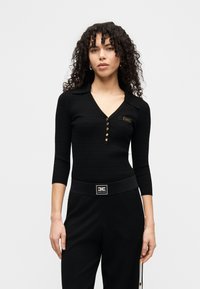 Μαύρο ριμπ μακρυμάνικο τοπ με V-λαιμόκοψη, που έχει χρυσά κουμπιά και μια μικρή ετικέτα με λογότυπο στο στήθος. Συνδυάζεται με μαύρο παντελόνι ψηλόμεσο.