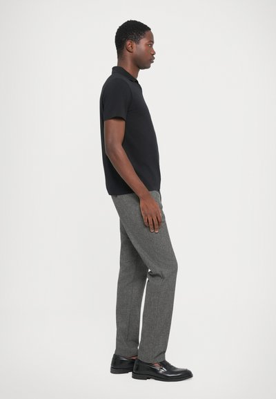 Schwarzes kurzärmeliges Polohemd, graue maßgeschneiderte Hose mit Hahnentrittmuster und schwarze Schuhe, von der Seite vor einem weißen Hintergrund betrachtet.