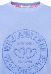 Jasnoniebieska bluza z okrągłym haftowanym logo, na którym widnieje napis „STAY WILD” oraz „EST 2014”, z dwoma wzorami serc i ribbowanym dekoltem.