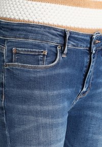 Vaqueros de mezclilla azul oscuro con cintura de altura media, que presentan un desgaste claro, cinco bolsillos y costuras en contraste. Textura lisa con un corte ajustado.