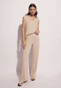 Beżowy, bezrękawnikowy kombinezon z dekoltem w kształcie litery V i elastycznym pasem. Połączony z czarnymi sandałami z falbaniastymi akcentami. Gładka tekstura.