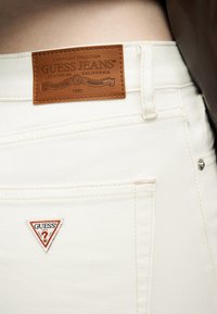 Κοντινό πλάνο άσπρων τζιν Guess που δείχνει το καφέ δερμάτινο ετικέτα της μάρκας κοντά στη μέση και το τρίγωνο κόκκινο και λευκό λογότυπο στην πίσω τσέπη.