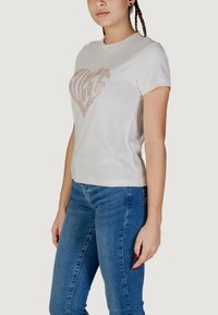 T-shirt bianco in cotone a maniche corte. Presenta un design di cuore scintillante di colore rosa chiaro sulla parte anteriore. Abbinato a jeans in denim blu.