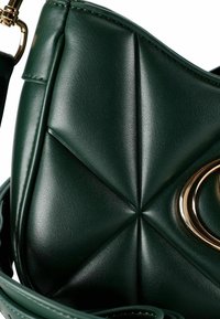 Bolso de piel verde oscuro con costura geométrica, que cuenta con un detalle de anillo de metal dorado y un clip de correa desmontable.