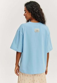 Světle modré oversized tričko vyrobené z měkké bavlny, s hranatou nálepkou na zadní straně a designem slunce, v uvolněném střihu.