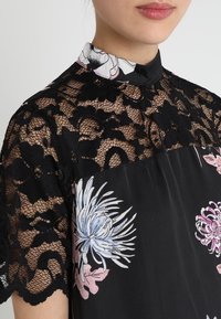 Top de encaje negro con patrón floral; presenta un cuello alto y mangas cortas, mostrando una intrincada textura de encaje y coloridos acentos florales.