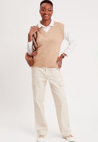 Un chaleco de punto marrón sobre una camisa blanca con cuello, combinado con pantalones cargo beige. El conjunto incluye un bolso de hombro con una correa estampada.