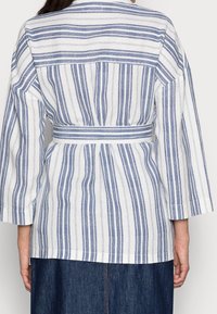 Valkoinen ja siniraitainen paita, jossa on kuminauhalla kiristetty vyötärö, kolmanneksen pituiset hihat ja rento istuvuus. Rakeinen puuvillakangas.