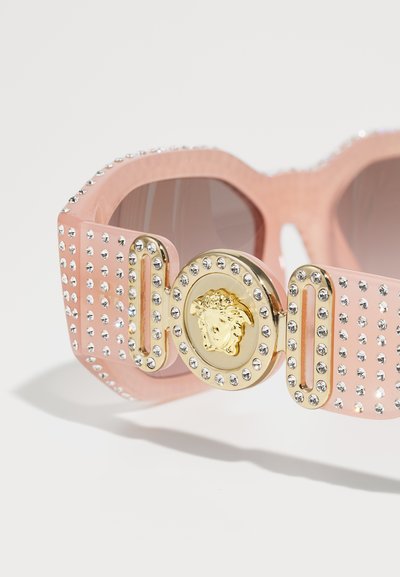 Óculos de sol rosa com um design geométrico robusto, adornados com strass transparentes e um detalhe redondo dourado com um motivo de leão.