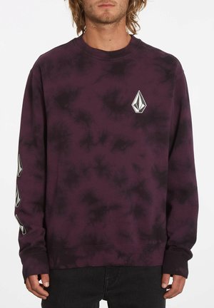 Mann trägt ein dunkelviolettes und schwarzes Batik-Sweatshirt mit geometrischen Logos auf der Brust und dem linken Ärmel, kombiniert mit schwarzen Hosen.