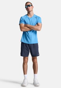 Heled sinine sportlik särk lühikeste varrukatega, millele on sobitatud tumesinised lühikesed püksid. Kanda prille ja valgeid tennisetossusid; käed ristis, seistes lihtsal taustal.