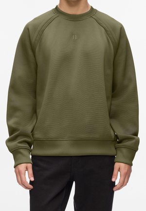 Person trägt ein olivgrünes Sweatshirt mit Rundhalsausschnitt, gerippten Bündchen und Saum, kombiniert mit einer schwarzen Hose, stehend vor weißem Hintergrund.