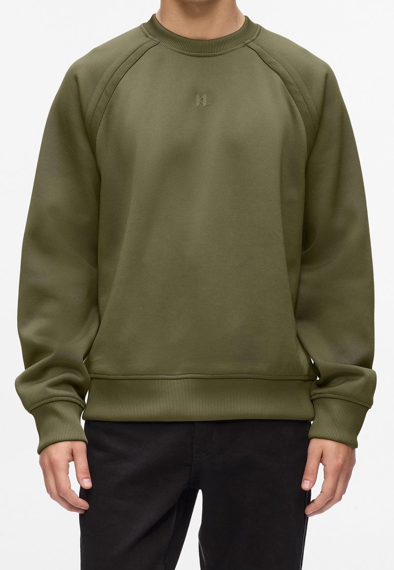 Une personne porte un sweat-shirt vert olive à col rond avec des poignets et un ourlet côtelés, associé à un pantalon noir, debout devant un fond blanc.