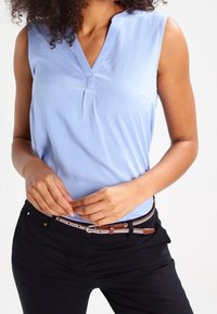 Ljusblå ärmlös blus med V-ringning och veckdetaljer. Kombinerad med svarta byxor och ett brunt bälte med en silverspänne.