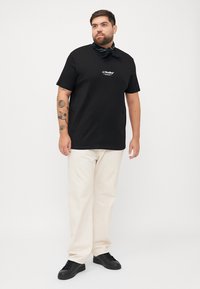 Schwarzes Baumwoll-T-Shirt mit Logo auf der Brust, kombiniert mit cremefarbenen Hosen und schwarzen Sneakers. Accessoire: gemusterte schwarze Bandana um den Hals.